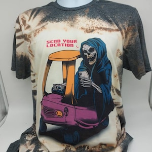 May include: A gray tie-dye t-shirt with a bleached design featuring a skeleton wearing a hooded robe and driving a pink toy car. The text "SEND YOUR LOCATION" is printed on the shirt.
