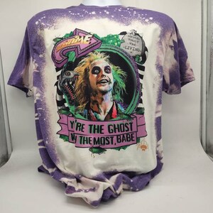 May include: A white t-shirt with a purple bleach dye effect. The shirt features a graphic of a character from the movie "Beetlejuice" with the text "Never trust the living" and "You're the ghost with the most, babe".