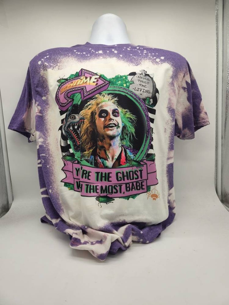 May include: A white t-shirt with a purple bleach dye effect. The shirt features a graphic of a character from the movie "Beetlejuice" with the text "Never trust the living" and "You're the ghost with the most, babe".