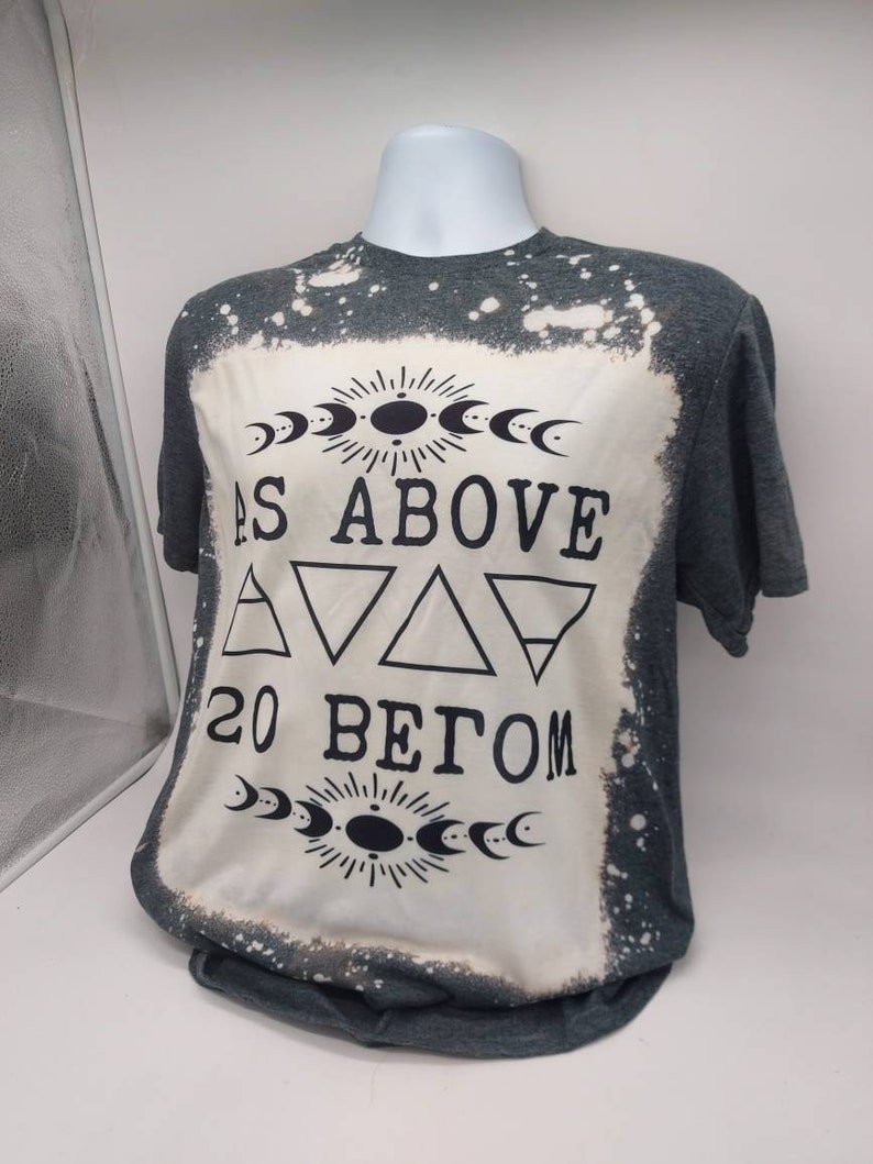 May include: A gray t-shirt with a bleached design featuring the text "As Above So Below" with a sun and moon symbol above and below the text.