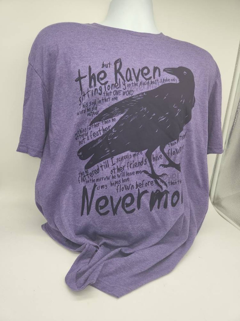 Op de afbeelding: Paars T-shirt met een zwart silhouet van een raaf en de tekst "the Raven sitting lonely on the placid bust spoke only this one word nevermore".