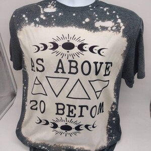 May include: A gray t-shirt with a bleach-splattered design. The shirt features a black graphic with the text "As Above So Below" and a symbol with three triangles.