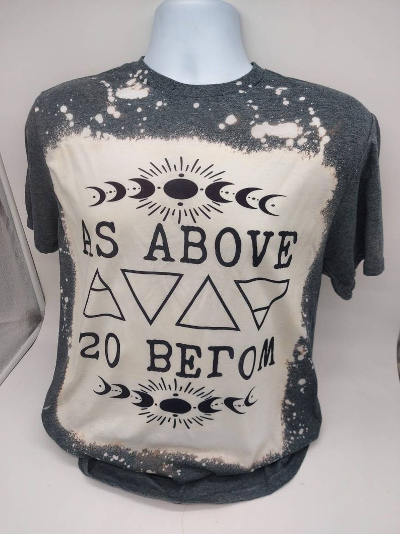 May include: A gray t-shirt with a bleach-splattered design. The shirt features a black graphic with the text "As Above So Below" and a symbol with three triangles.