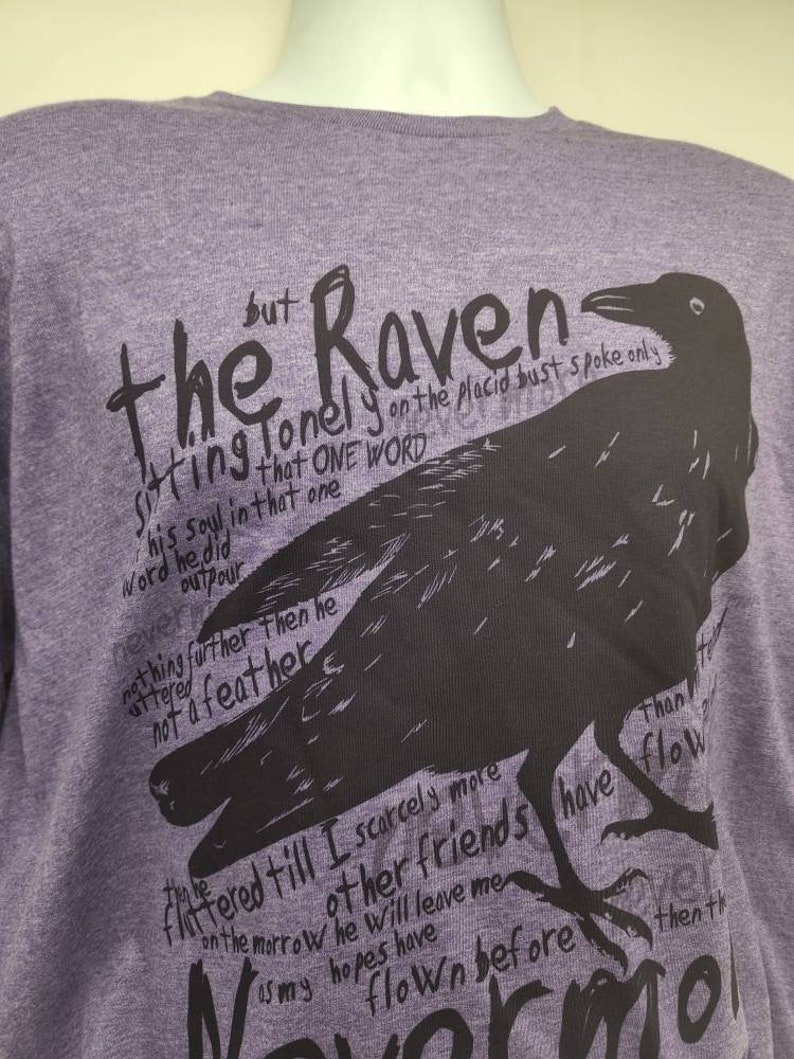 Op de afbeelding: Paars T-shirt met een zwarte raaf afbeelding en tekst die luidt "The Raven sitting lonely on the placid bust spoke only his soul in that one word he did outpour nothing further then he uttered not a feather fluttered till I scarcely more on the morrow other friends have flown before than my hopes have flown then the"