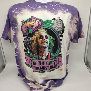 May include: A white t-shirt with a purple bleach dye effect. The shirt features a graphic of a character from the movie Beetlejuice. The graphic includes the text "You're the ghost with the most, babe" and "Never trust the living".