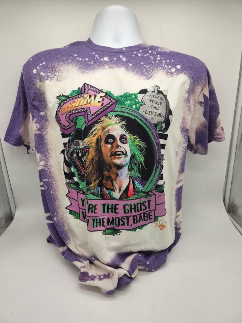 May include: A white t-shirt with a purple bleach dye effect. The shirt features a graphic of a character from the movie Beetlejuice. The graphic includes the text "You're the ghost with the most, babe" and "Never trust the living".