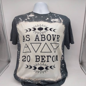 May include: A gray t-shirt with a bleach-splattered design. The shirt features a black graphic with the text "As Above So Below" and a symbol with triangles and moons.