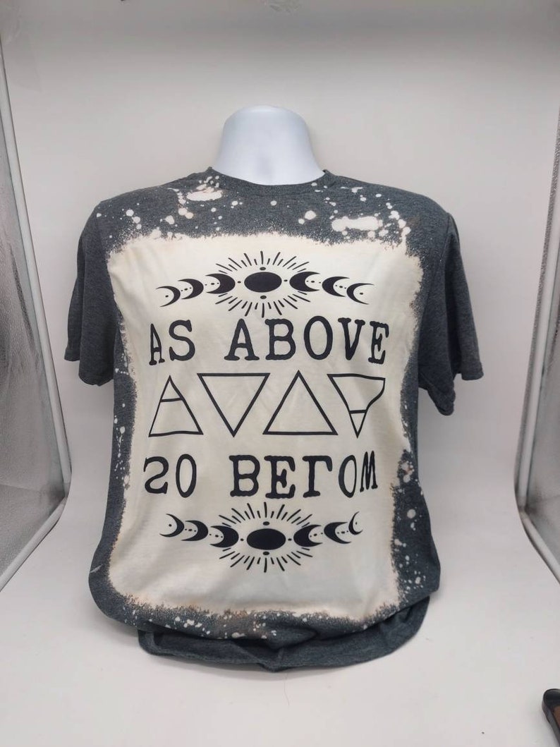 May include: A gray t-shirt with a bleach-splattered design. The shirt features a black graphic with the text "As Above So Below" and a symbol with triangles and moons.