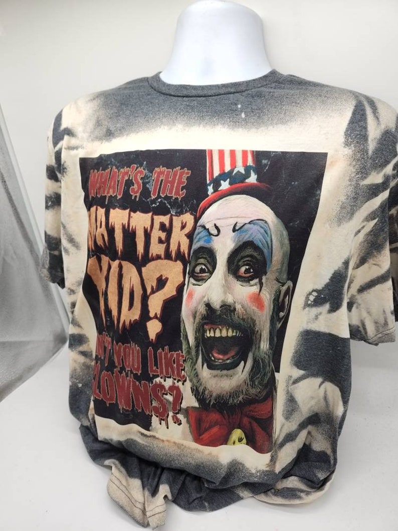 May include: A grey t-shirt with a bleach-splattered design featuring a graphic of a clown with a red, white, and blue hat. The graphic includes the text "What's the Matter Kid? Ain't You Like Clowns?"