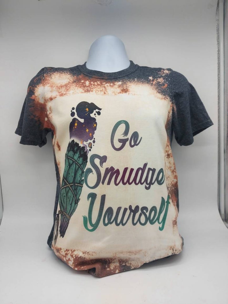 May include: A bleached grey t-shirt with a white design featuring a smudge stick and the text "Go Smudge Yourself".