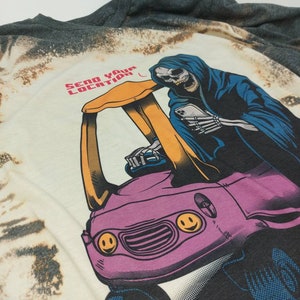 May include: A white t-shirt with a bleached effect featuring a graphic of a skeleton in a blue hooded robe driving a pink car with smiley faces. The text "SEND YOUR LOCATION" is printed in red and black on the shirt.