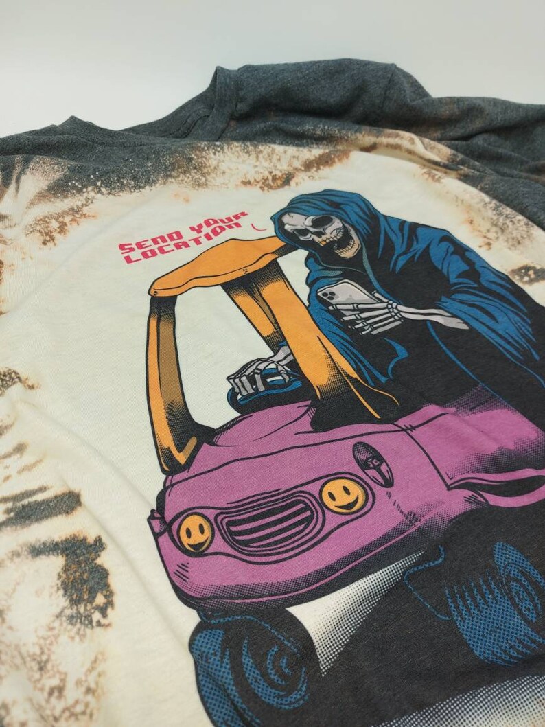 May include: A white t-shirt with a bleached effect featuring a graphic of a skeleton in a blue hooded robe driving a pink car with smiley faces. The text "SEND YOUR LOCATION" is printed in red and black on the shirt.