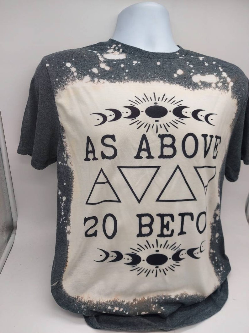 May include: A gray t-shirt with a bleached design featuring the text "As Above So Below" with a sun and moon symbol above and below the text. The design also includes three triangles.