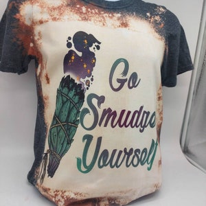 May include: A bleached grey t-shirt with a white design featuring a smudge stick and the text "Go Smudge Yourself".