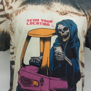 May include: A white t-shirt with a tie-dye pattern featuring a cartoon image of the Grim Reaper driving a small pink car with a yellow roof. The Grim Reaper is wearing a blue hooded robe and holding a smartphone. The text "SEND YOUR LOCATION" is printed above the image.