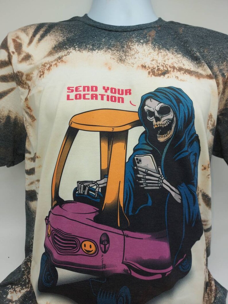 May include: A white t-shirt with a tie-dye pattern featuring a cartoon image of the Grim Reaper driving a small pink car with a yellow roof. The Grim Reaper is wearing a blue hooded robe and holding a smartphone. The text "SEND YOUR LOCATION" is printed above the image.