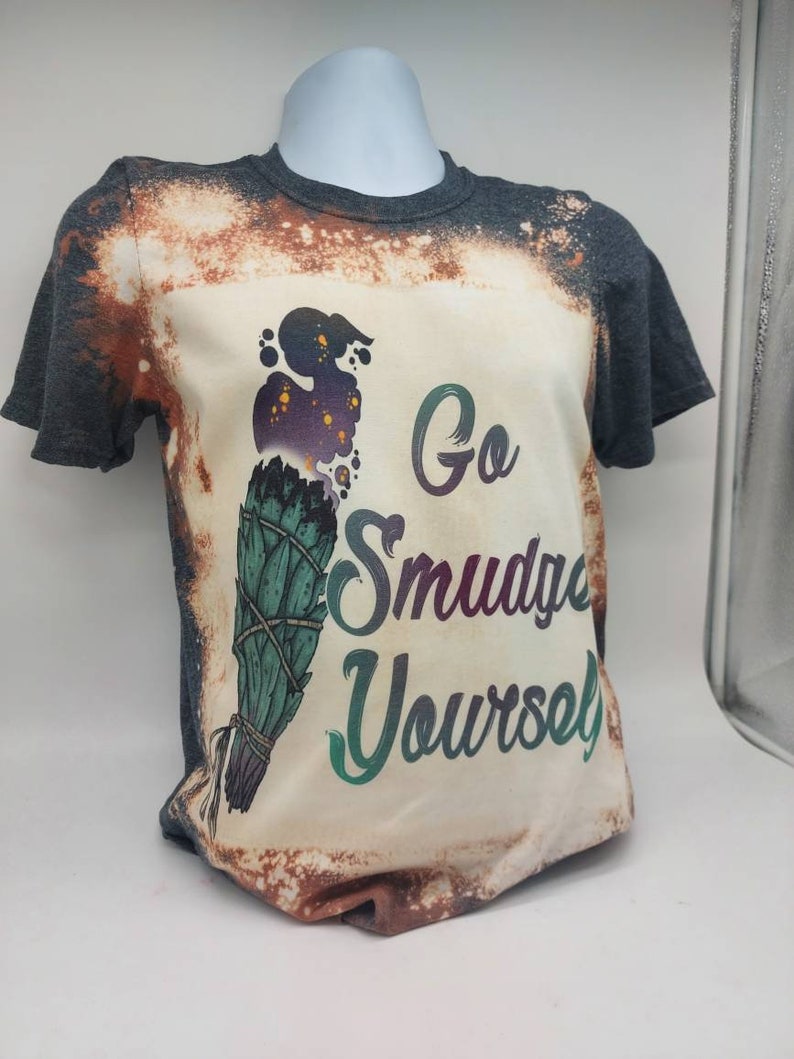 May include: A grey t-shirt with a bleached design featuring a sage smudge stick and the text "Go Smudge Yourself".