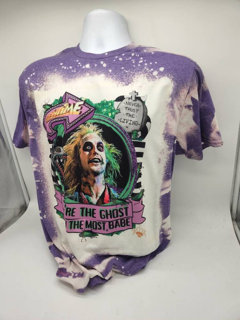 May include: Purple tie-dye t-shirt with a bleached effect. The shirt features a graphic of a character from the movie Beetlejuice, with the text "Be the Ghost with the Most, Babe" and "Never Trust the Living".