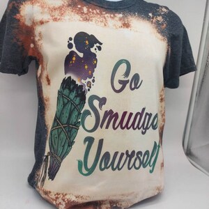 May include: A bleached grey t-shirt with a white design featuring a smudge stick and the text "Go Smudge Yourself".