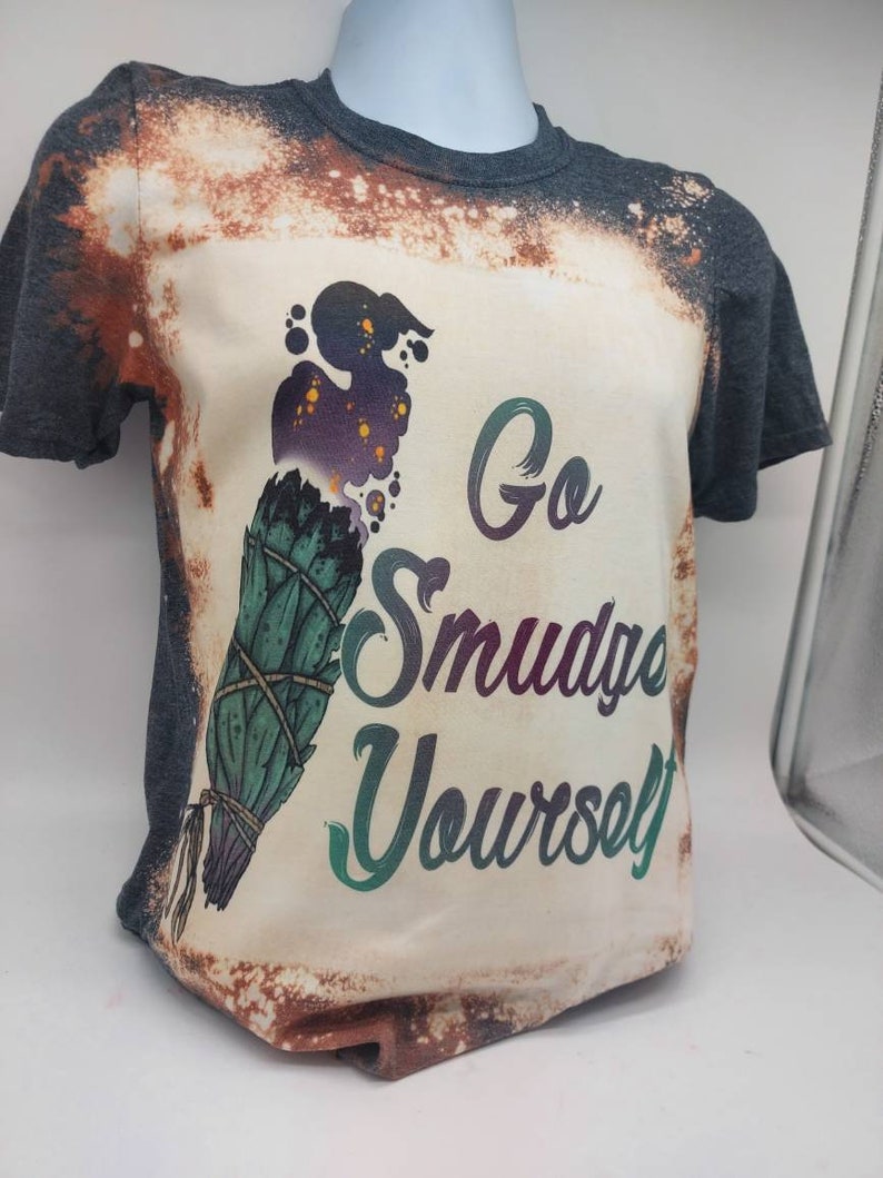May include: A bleached grey t-shirt with a white design featuring a smudge stick and the text "Go Smudge Yourself".