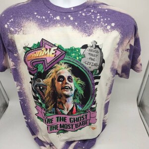 May include: A purple and white bleached t-shirt with a graphic of a character from the movie Beetlejuice. The graphic includes the text "Beetlejuice" and "You're the Ghost, the Most, Babe".