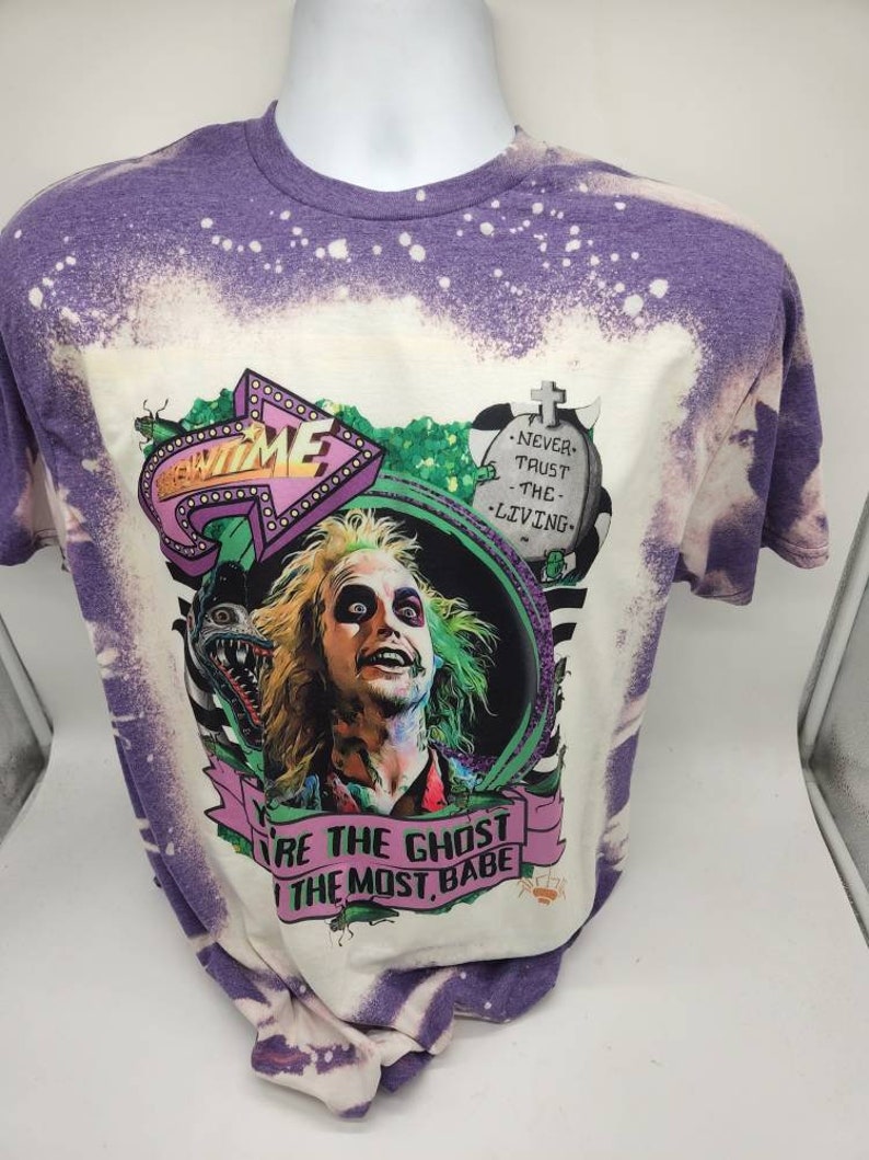 May include: A purple and white bleached t-shirt with a graphic of a character from the movie Beetlejuice. The graphic includes the text "Beetlejuice" and "You're the Ghost, the Most, Babe".