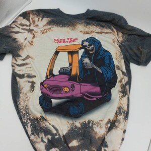 May include: A gray t-shirt with a bleach-splattered design featuring a cartoon skeleton driving a pink toy car. The skeleton is wearing a blue hooded robe and holding a smartphone. The text "Send Your Location" is printed above the car.