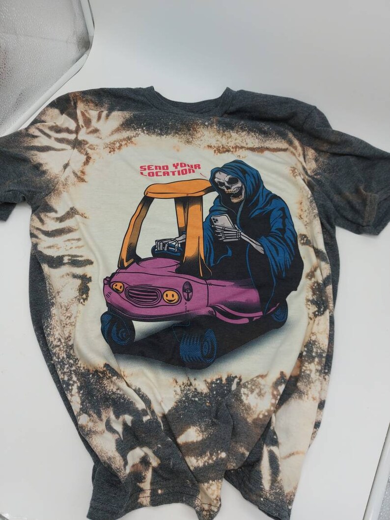 May include: A gray t-shirt with a bleach-splattered design featuring a cartoon skeleton driving a pink toy car. The skeleton is wearing a blue hooded robe and holding a smartphone. The text "Send Your Location" is printed above the car.