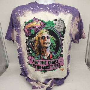 May include: A white and purple tie-dye t-shirt with a graphic print of a character from the movie Beetlejuice. The graphic features the character's face and a green and black background. The text "You're the ghost with the most, babe" is printed on the shirt. The text "Never trust the living" is printed on the shirt.