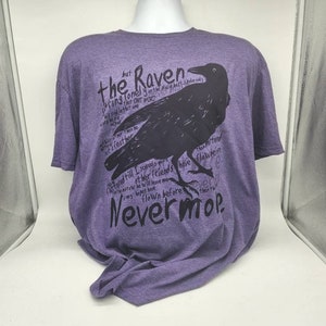 Op de afbeelding: Paars T-shirt met een zwarte raaf afbeelding en tekst die luidt "The Raven sitting lonely on the placid bust spoke only that one word... Nevermore."