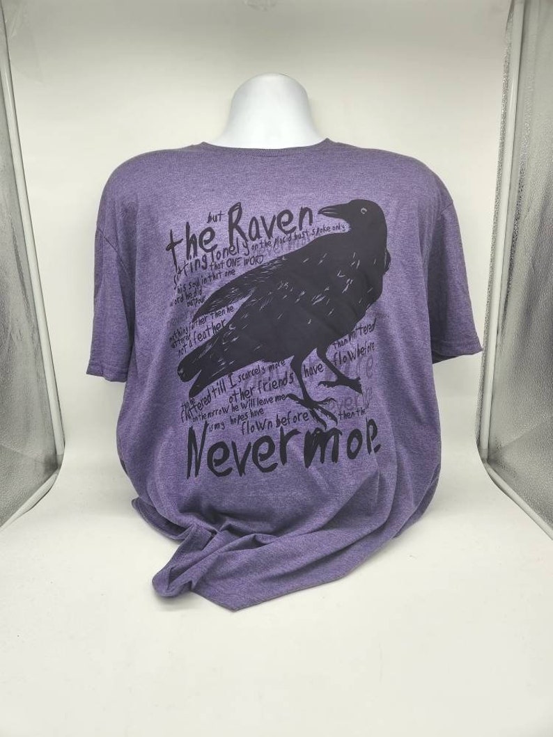 Op de afbeelding: Paars T-shirt met een zwarte raaf afbeelding en tekst die luidt "The Raven sitting lonely on the placid bust spoke only that one word... Nevermore."