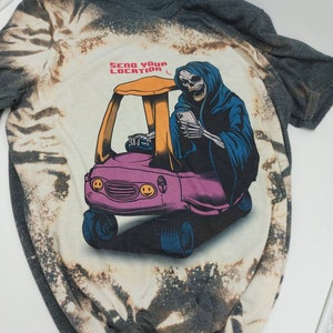 May include: A grey t-shirt with a bleach-splattered design featuring a cartoon Grim Reaper driving a toy car. The Grim Reaper is holding a smartphone and the text "SEND YOUR LOCATION" is above the car.
