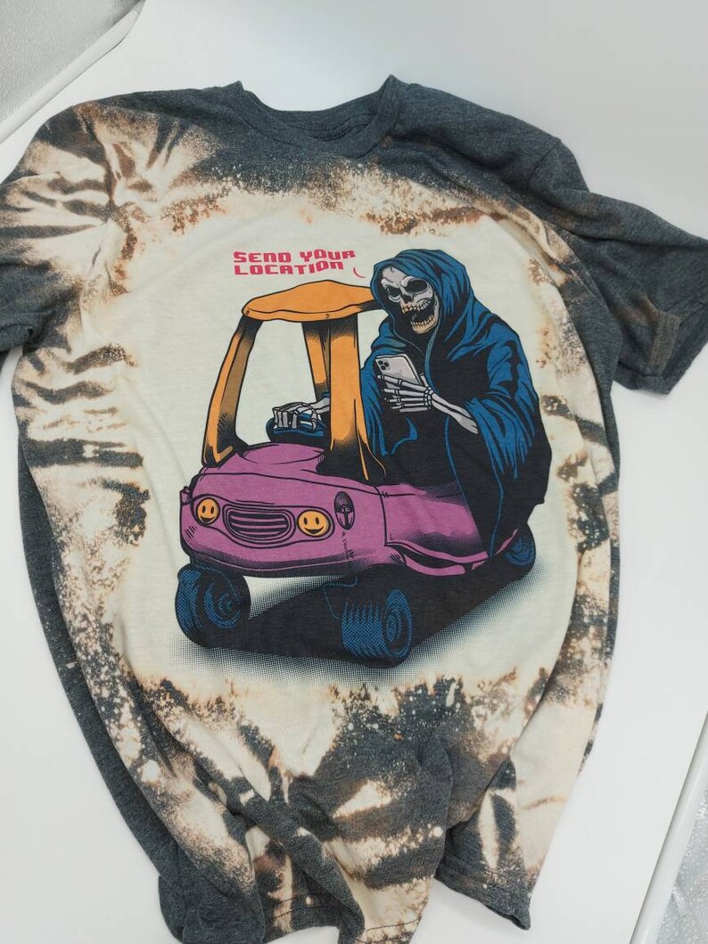 May include: A grey t-shirt with a bleach-splattered design featuring a cartoon Grim Reaper driving a toy car. The Grim Reaper is holding a smartphone and the text "SEND YOUR LOCATION" is above the car.