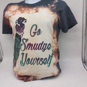 May include: A grey t-shirt with a white bleached design. The design features a green and purple smudge stick with the text "Go Smudge Yourself".