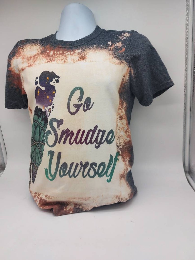May include: A grey t-shirt with a white bleached design. The design features a green and purple smudge stick with the text "Go Smudge Yourself".