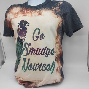 May include: A grey t-shirt with a bleached effect and a graphic of a smudge stick with the text "Go Smudge Yourself".