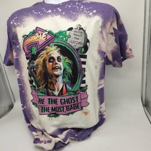 May include: Purple and white bleached t-shirt with a graphic print of a character from the movie Beetlejuice. The graphic includes the text "Be the ghost the most, babe" and "Never trust the living".