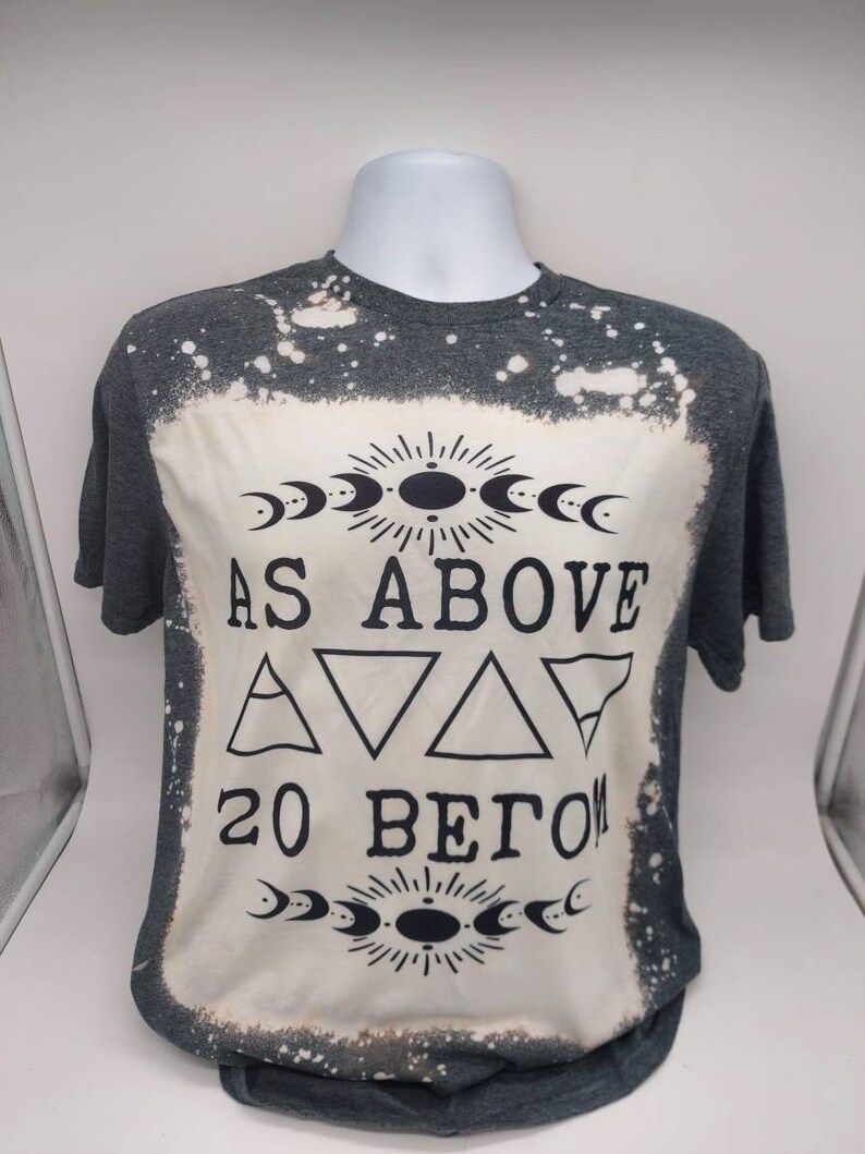 May include: A gray t-shirt with a bleach-splattered design. The shirt features a black graphic with the text "As Above So Below" and a symbol with three triangles.