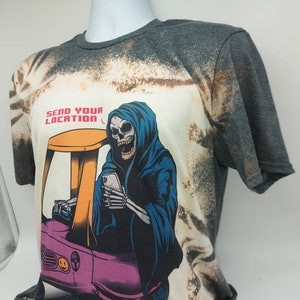 May include: A bleached grey t-shirt with a graphic of a skeleton in a blue hooded robe driving a small pink car. The skeleton is holding a smartphone and the text "Send Your Location" is printed above the graphic.