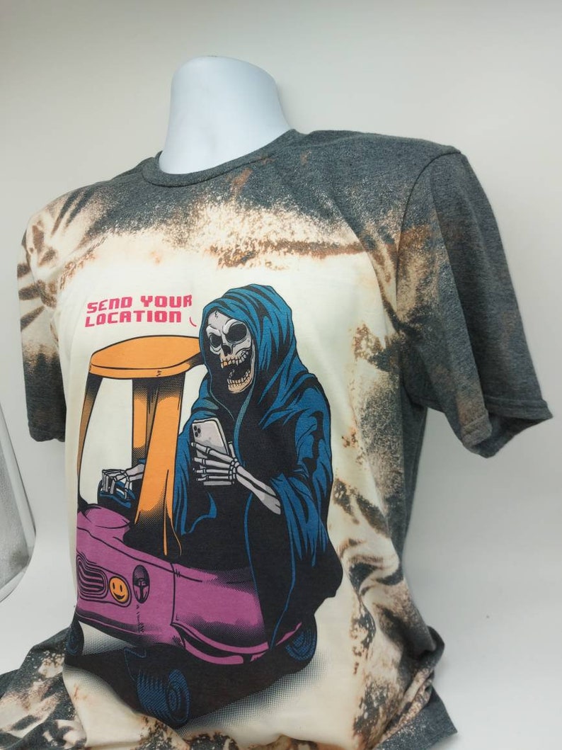 May include: A bleached grey t-shirt with a graphic of a skeleton in a blue hooded robe driving a small pink car. The skeleton is holding a smartphone and the text "Send Your Location" is printed above the graphic.