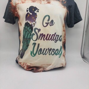 May include: A bleached grey t-shirt with a white design featuring a green and purple smudge stick with the text "Go Smudge Yourself".