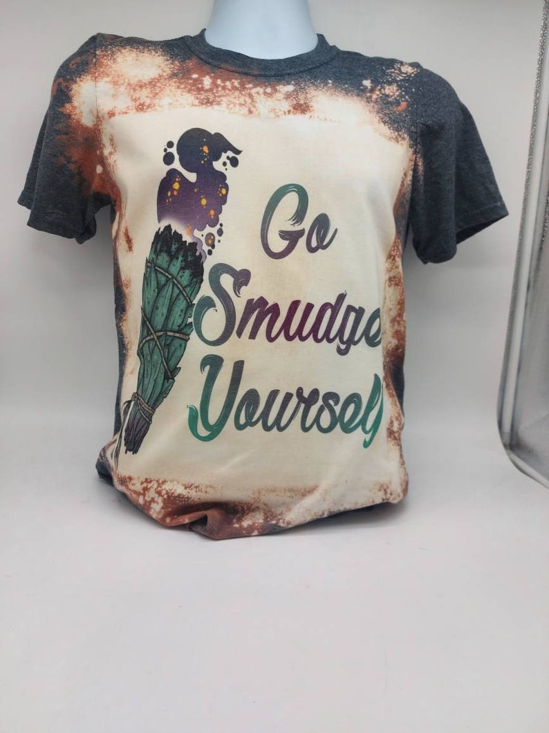 May include: A bleached grey t-shirt with a white design featuring a green and purple smudge stick with the text "Go Smudge Yourself".