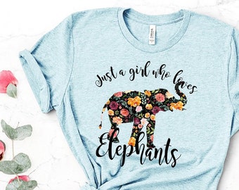 May include: A light blue heathered t-shirt featuring a black graphic of an elephant filled with a colorful floral pattern. The text "Just a girl who loves Elephants" is printed in black script above and below the elephant silhouette.
