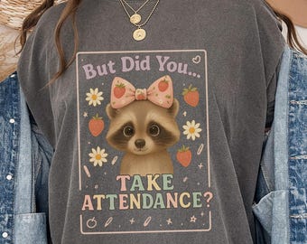 May include: Dark gray t-shirt featuring a raccoon with a pink bow, surrounded by daisies and strawberries. The text reads "But Did You... TAKE ATTENDANCE?" in a playful font. The shirt is part of a casual wear collection.