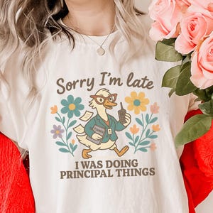 May include: A cream-coloured t-shirt featuring the text "Sorry I'm late" above a cartoon duck wearing glasses and holding a planner and a phone. The duck is surrounded by flowers, with the text "I was doing principal things" below.