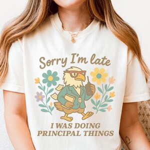 May include: A cream-coloured t-shirt featuring the text "Sorry I'm late" above a cartoon eagle wearing glasses and holding a phone. The eagle is surrounded by flowers, with the text "I WAS DOING PRINCIPAL THINGS" below.