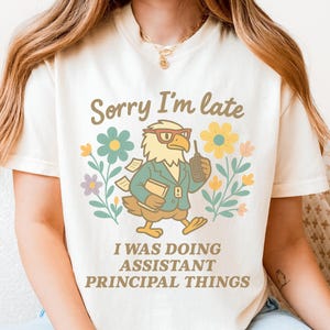 May include: A cream-coloured t-shirt featuring the text "Sorry I'm late" above a cartoon eagle wearing glasses and holding a phone. The eagle is surrounded by flowers and the text "I WAS DOING ASSISTANT PRINCIPAL THINGS".