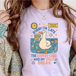 May include: Lilac t-shirt featuring a cartoon duck with a coffee cup, text reads "Sorry I'm Late" and "The Coffee Line Was My Only Break." The design includes a clock, lockers, and floral accents. A Teacher Lounge sign is also visible.