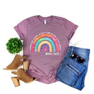 May include: A mauve short-sleeve t-shirt featuring a colourful rainbow graphic with the text Teach Love Inspire Motivate Lead Encourage Listen Connect and #MISS KATY, paired with blue denim jeans, tan perforated block-heel sandals, and sunglasses.