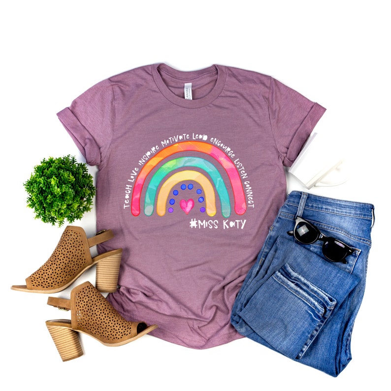 May include: A mauve short-sleeve t-shirt featuring a colourful rainbow graphic with the text Teach Love Inspire Motivate Lead Encourage Listen Connect and #MISS KATY, paired with blue denim jeans, tan perforated block-heel sandals, and sunglasses.